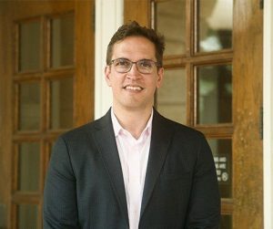 Image of a white man standing in front of a wooden door wearing a dress shirt, jacket ,and black glasses.