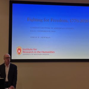 A speaker stands at a podium beside a projected slide titled “Fighting for Freedom, 1776–2026”