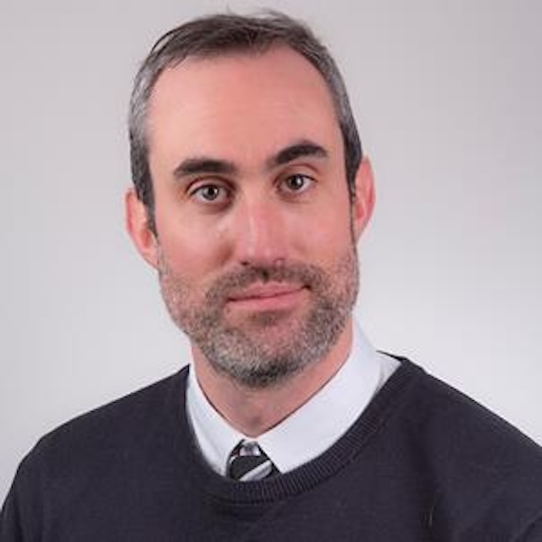 Headshot of a man with short brown hair and a beard, wearing a collared shirt, tie, and black sweater.