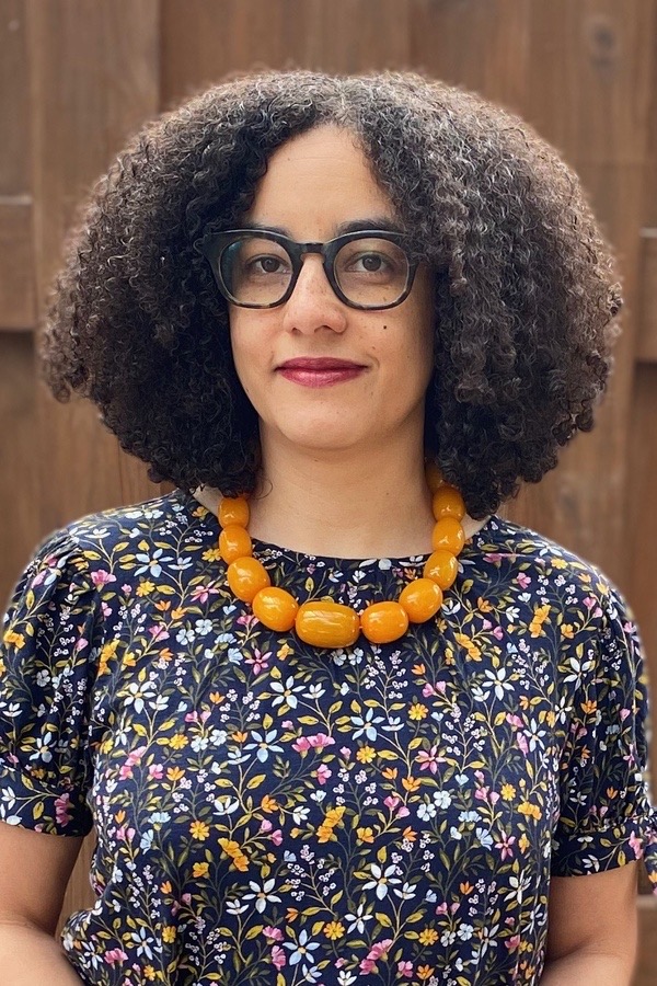 A medium-skin-toned woman with dark, coily hair faces the camera. She is wearing dark brown glasses, a dark blue top with small orange, pink, and blue flowers, and a neckless with large orange beads.