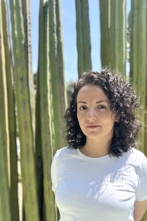 Picture of a woman with dark curly hair standing outside with tall cacti behind her.