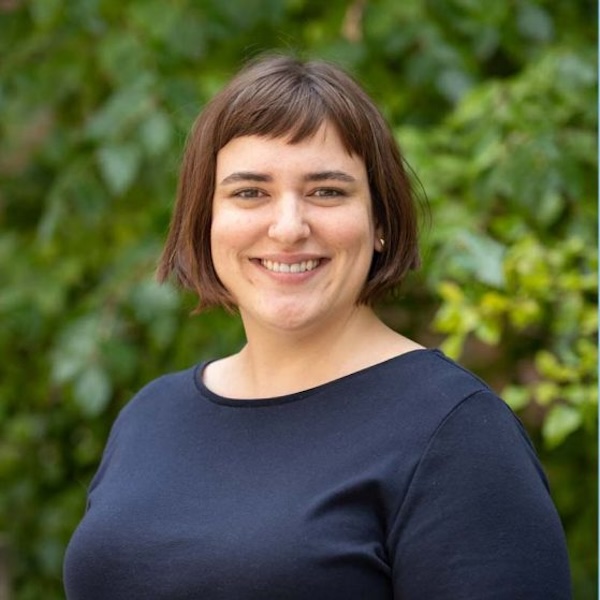 This is a headshot of a woman with short brown hair standing outside with greenery behind her.