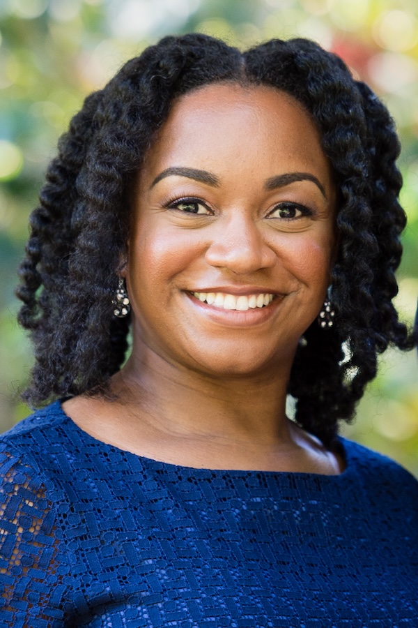 This is an image of a woman with tan skin, thick curly hair, and wearing a navy blue top smiling.