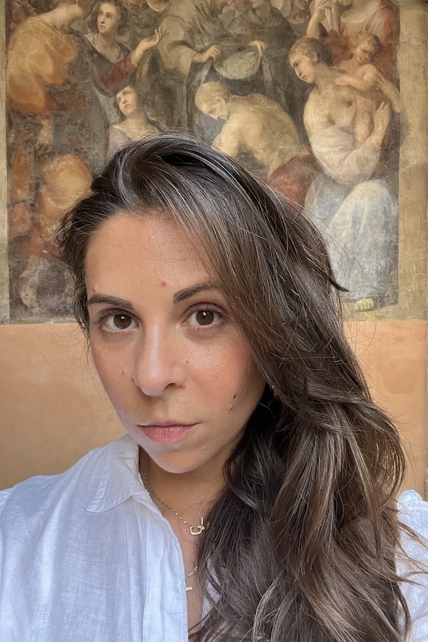 This is a headshot of a woman with long brown hair standing in front of Renaissance fresco.