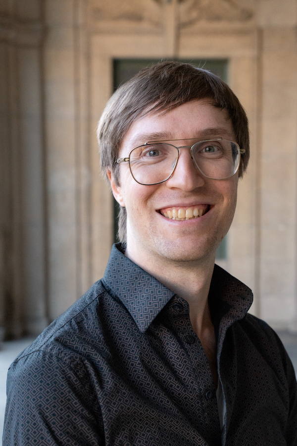 This is a headshot of a man with brown hair and wire-frame glasses standing in front of a yellow standstone wall.