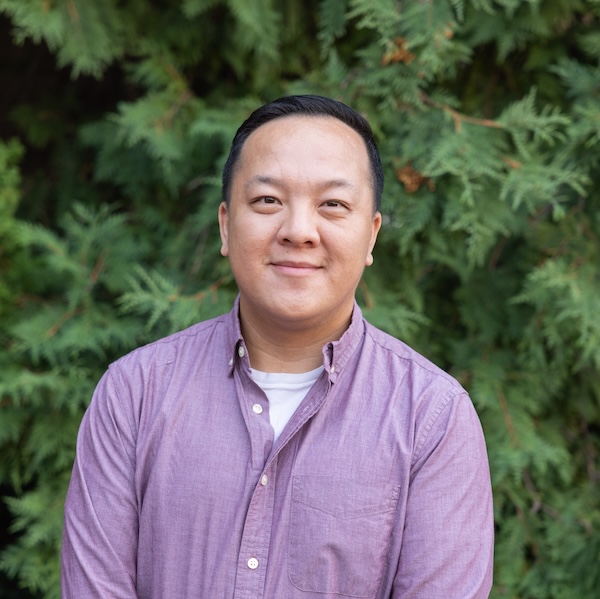 Image of a band standing in front of greenery. He has tan skin, short black hair, and is wearing a purple button down shirt.