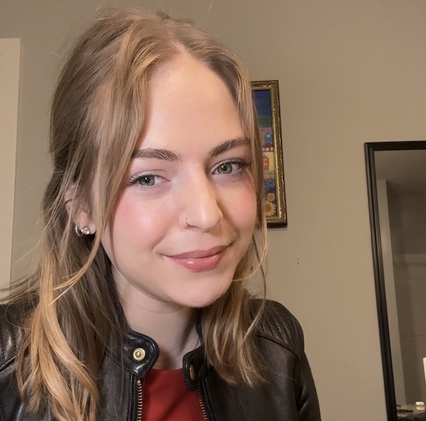 Image of a woman with blonde hair and fair skin wearing a leather jacket and red shirt, smiling. 