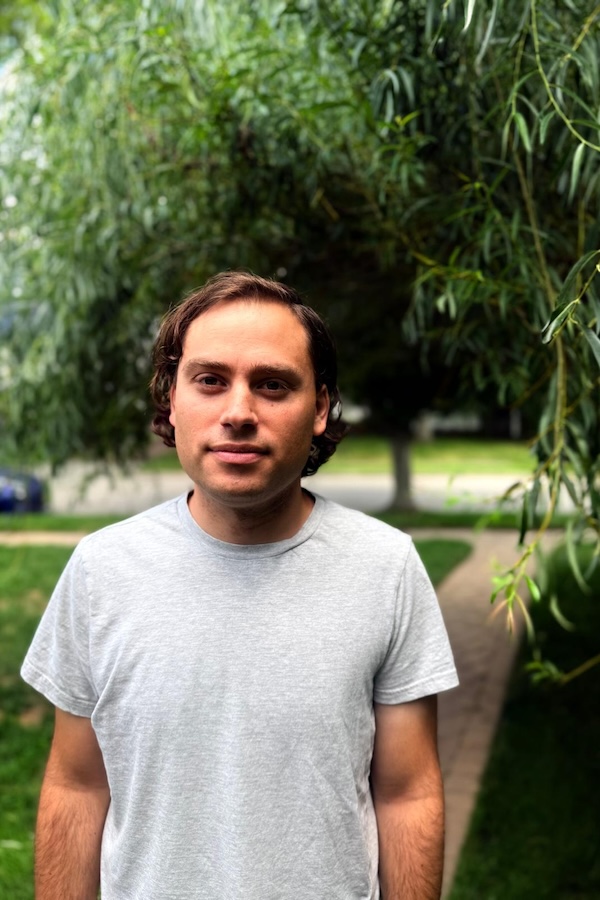 This is a headshot of a man wearing a white t-shirt standing outside with a tree in the background.