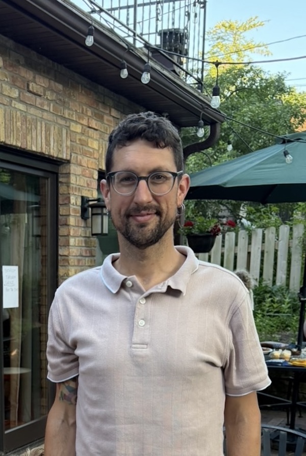 Image of a person standing in front of a brick building wearing a polo shirt and glasses.