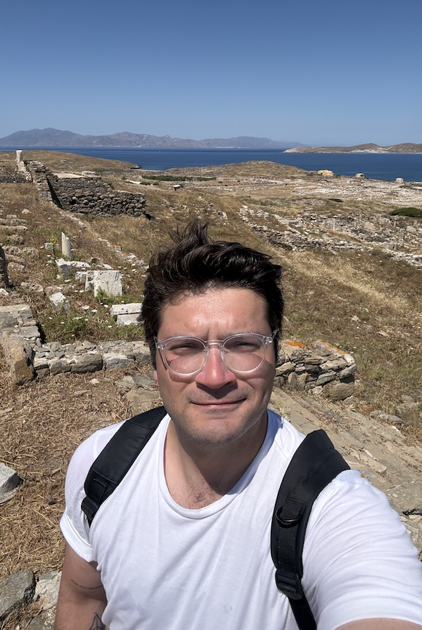 A person wearing a white T-shirt, glasses, and a backpack takes a selfie at an ancient archaeological site overlooking the sea, with stone ruins, small islands, and distant mountains under a clear blue sky.