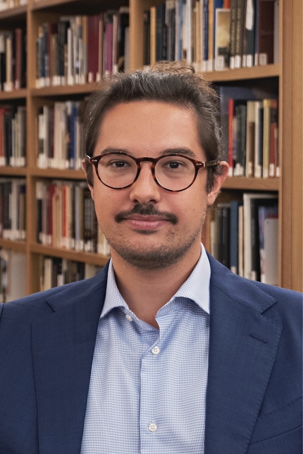 This is a headshot of a man wearing glasses, a blue suit, and a blue collar shirt with a library on the background.